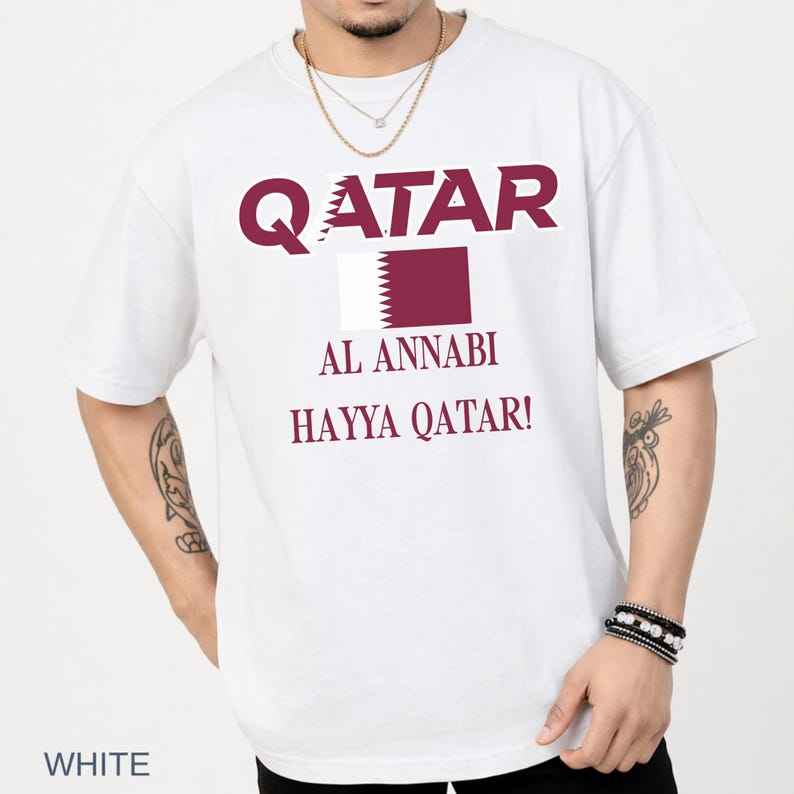 A male model in a crisp white t-shirt showcasing the maroon Qatar football design, styled with layered necklaces and beaded wrist bracelets in a clean, well-lit lifestyle studio.