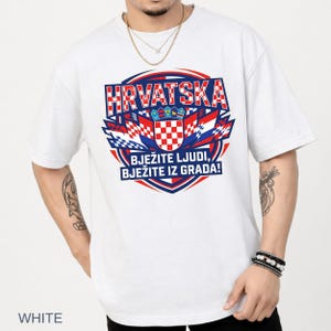 A tattooed male model wearing a clean white t-shirt that displays a red, blue, and white Croatian coat of arms crest emblem and fan chant text.