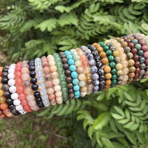 Peut inclure: Une collection de bracelets de perles de différentes couleurs, dont orange, blanc, noir, rose, bleu, vert et marron. Les bracelets sont présentés sur une surface cylindrique beige sur fond de feuillage vert. Chaque bracelet est composé de perles rondes.