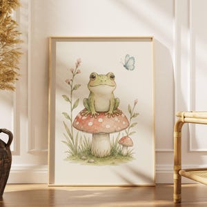 Pode incluir: Impressão artística em aquarela de um sapo verde sobre um cogumelo vermelho e branco. Uma borboleta azul e flores cor-de-rosa completam a cena. A obra, emoldurada em uma moldura dourada, evoca um ambiente de conto de fadas.
