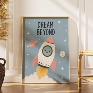Könnte beinhalten: Gerahmter Kunstdruck mit einer Cartoon-Rakete mit lächelndem Gesicht, die durch einen blauen Himmel mit Sternen und Planeten aufsteigt. Der Text "DREAM BEYOND" ist oben zu sehen. Die Rakete hat rosa Flossen und gelbe Flammen.