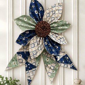 May include: A decorative fabric flower wall hanging featuring layered petals in navy blue, sage green, and cream with floral and geometric patterns. The center is a textured, dark brown circular element. The petals are arranged in a starburst shape.