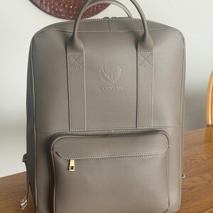 Könnte beinhalten: Ein taupefarbener Rucksack mit zwei Tragegriffen, einer vorderen Reißverschlusstasche und einem Hauptfach mit Reißverschluss. Auf der Vorderseite ist der Text CORVIAN sowie ein geflügeltes Logo eingeprägt. Das Design zeigt sichtbare weiße Nähte.
