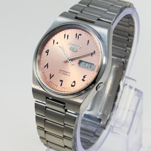 May include: A silver-toned Seiko 5 automatic wristwatch with a salmon-colored face. The watch features Arabic numerals, a day-date display, and a stainless steel band. The watch is displayed on a clear stand.