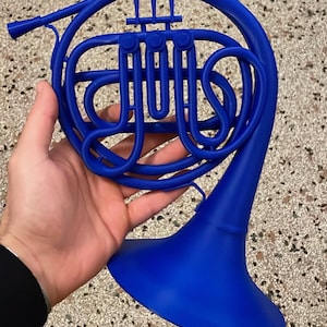 Op de afbeelding: Een helderblauwe Franse hoorn, een koperen blaasinstrument, wordt in een hand gehouden. De ingewikkelde buizen en de beker van de hoorn zijn zichtbaar, wat het opgerolde ontwerp benadrukt. Het instrument is een solide, uit één stuk bestaande constructie.
