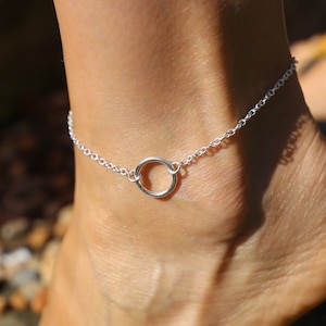 May include: A silver anklet with a circle charm. The anklet is on a person's ankle.