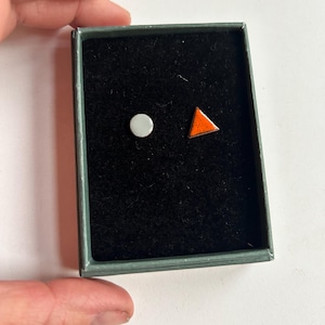 May include: A small box with a black velvet interior containing two ceramic earrings. One earring is a white circle and the other is an orange triangle.
