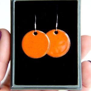May include: A pair of orange round ceramic earrings with silver hooks. The earrings are displayed in a black box.