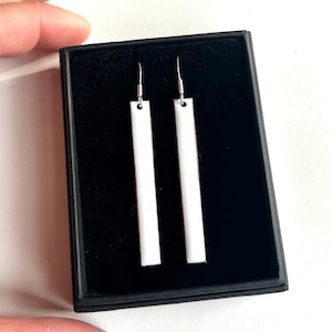 May include: Two white rectangular earrings with silver hooks, displayed in a black box. The earrings are long and slender, with a minimalist design. The earrings are presented against a white background.