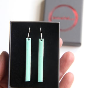 May include: A pair of light green ceramic earrings with silver hooks, displayed in a black box.