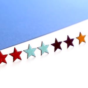 Könnte beinhalten: Eine Reihe von sieben emaillierten sternförmigen Pins in verschiedenen Farben, darunter Rot, Blau, Grün, Lila und Orange. Die Pins sind in einer Linie auf einer weißen Oberfläche angeordnet.