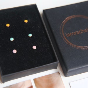 May include: A black box with a velvet interior containing five pairs of small, colourful stud earrings. The earrings are yellow, pink, and two shades of blue.