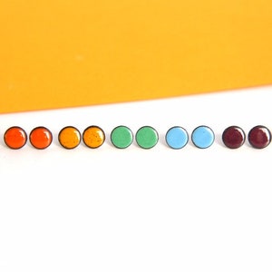 Könnte beinhalten: Ein Set mit zehn kleinen runden Ohrsteckern in verschiedenen Farben, darunter Rot, Orange, Grün, Blau, Lila und Weinrot. Die Ohrstecker haben einen schwarzen Rand.