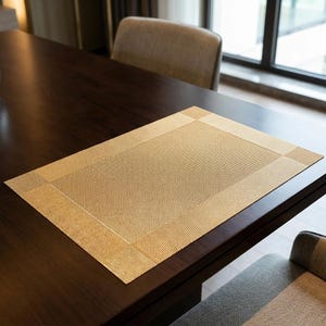 Op de afbeelding: Een rechthoekige, geweven placemat in een warme, goudbruine tint. De placemat heeft een gestructureerd oppervlak en een subtiel randontwerp. Het ligt op een donkerbruine houten tafel, wat wijst op een eet- of keukensetting.