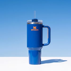 Pode incluir: Um copo Stanley azul com uma al&ccedil;a e uma palhinha transparente. O copo tem uma tampa prateada e o logotipo Stanley em laranja. O copo &eacute; cil&iacute;ndrico e assenta numa superf&iacute;cie branca sobre um fundo azul.