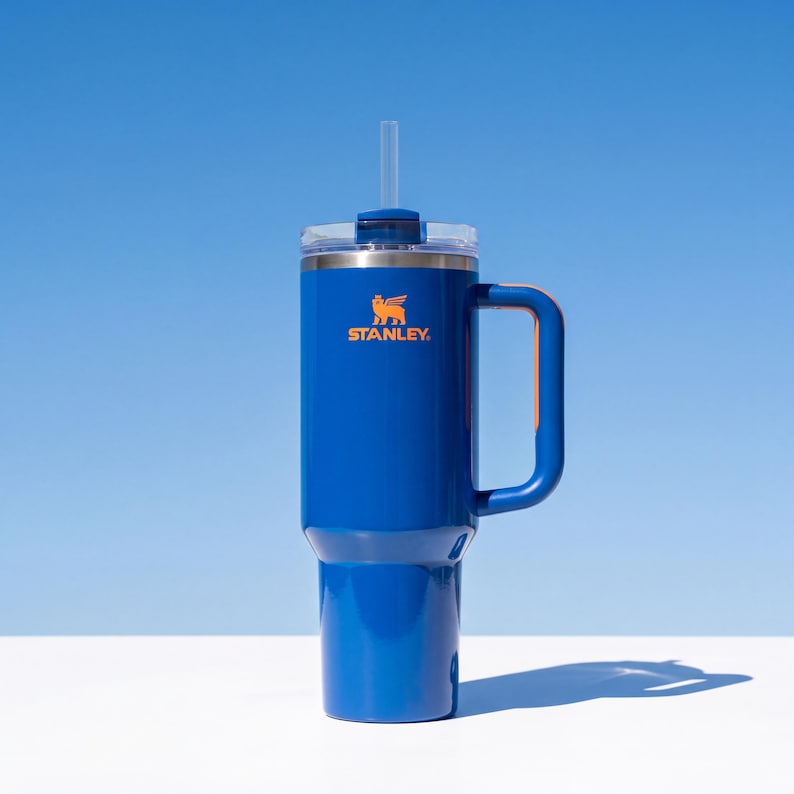 Pode incluir: Um copo Stanley azul com uma al&ccedil;a e uma palhinha transparente. O copo tem uma tampa prateada e o logotipo Stanley em laranja. O copo &eacute; cil&iacute;ndrico e assenta numa superf&iacute;cie branca sobre um fundo azul.