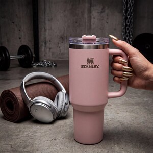 May include: A pink Stanley travel mug with a handle and a clear lid. The Stanley logo is visible on the front. The mug is held by a person with gold-colored nails. A rolled-up yoga mat and headphones are in the background.