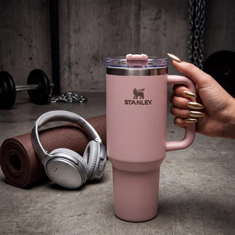 May include: A pink Stanley travel mug with a handle and a clear lid. The Stanley logo is visible on the front. The mug is held by a person with gold-colored nails. A rolled-up yoga mat and headphones are in the background.