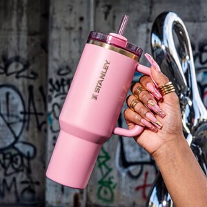 May include: A pink Stanley tumbler with a handle and straw. The tumbler has a gold rim and the Stanley logo. The tumbler is held by a hand with long, decorated nails and gold rings. The background is a concrete wall with graffiti.
