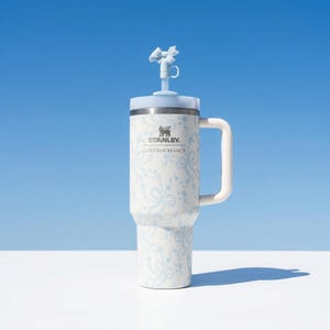 May include: A white and light blue Stanley tumbler with a handle and a decorative bow on the lid. The tumbler has a floral pattern and the text "STANLEY LOVESHACKFANCY". The background is a gradient of blue and white.