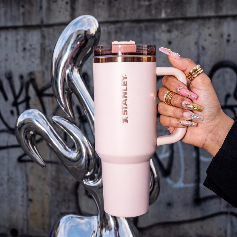 May include: A light pink Stanley travel mug with a handle and a clear lid. The mug has a gold rim and the Stanley logo printed on the side. The mug is held by a person with long, decorated nails.