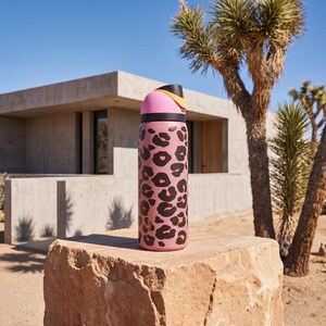 May include: A pink water bottle featuring a black and orange leopard print design. The bottle has a pink lid with black and yellow detailing. The bottle is set on a rock in an outdoor environment.