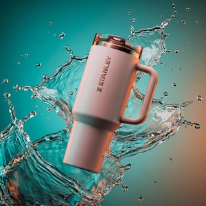 May include: A light pink Stanley tumbler with a handle and a lid, set against a backdrop of water splashing. The tumbler has the Stanley logo printed on the side. The background transitions from teal to orange.