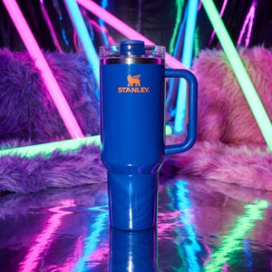 Pode incluir: Um copo Stanley azul com uma al&ccedil;a e uma tampa transparente. O logotipo da Stanley &eacute; impresso em laranja. O copo &eacute; cil&iacute;ndrico e parece ser feito de a&ccedil;o inoxid&aacute;vel. O fundo apresenta luzes de n&eacute;on e uma superf&iacute;cie rosa difusa.