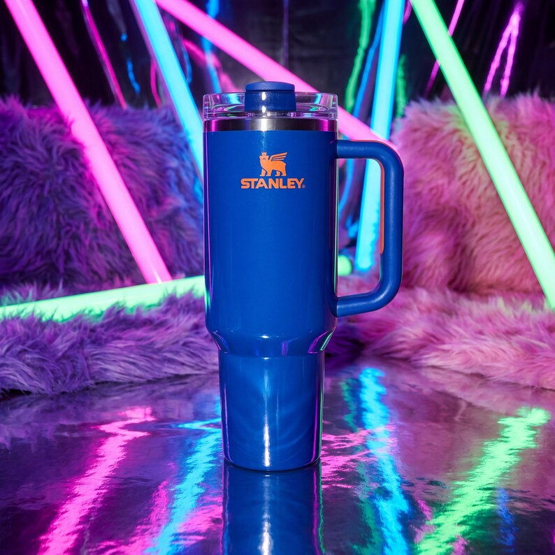 Pode incluir: Um copo Stanley azul com uma al&ccedil;a e uma tampa transparente. O logotipo da Stanley &eacute; impresso em laranja. O copo &eacute; cil&iacute;ndrico e parece ser feito de a&ccedil;o inoxid&aacute;vel. O fundo apresenta luzes de n&eacute;on e uma superf&iacute;cie rosa difusa.