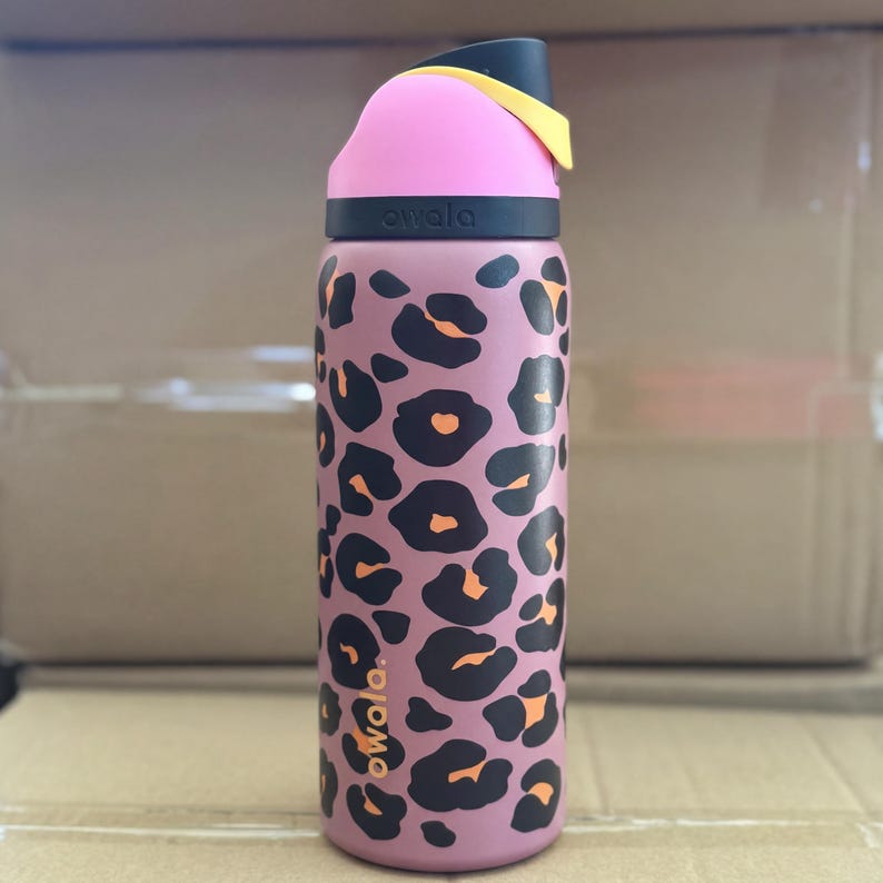 May include: A pink and black Owala water bottle with a leopard print pattern. The bottle features a black lid, a pink top, and yellow accents. The Owala logo is printed on the bottle.