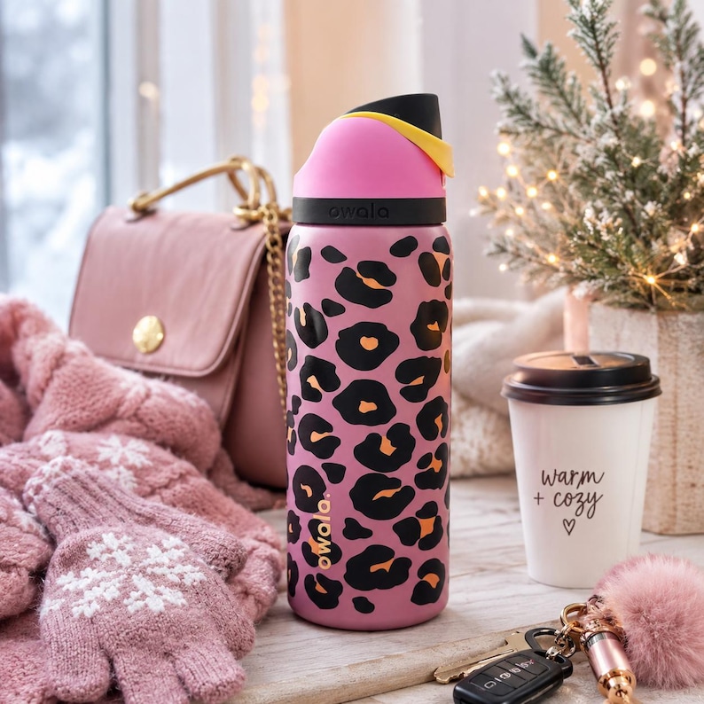 May include: A pink Owala water bottle with a black and orange leopard print. The bottle has a pink lid with a black and yellow handle. The image also includes a pink handbag, gloves, and a coffee cup with the text "warm + cozy".