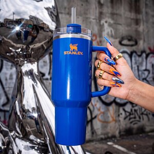 Pode incluir: Um copo Stanley azul com al&ccedil;a e canudo. O logotipo da Stanley &eacute; laranja. O copo &eacute; segurado em frente a uma parede coberta de grafite. A pessoa que segura o copo tem unhas longas e coloridas e an&eacute;is dourados.