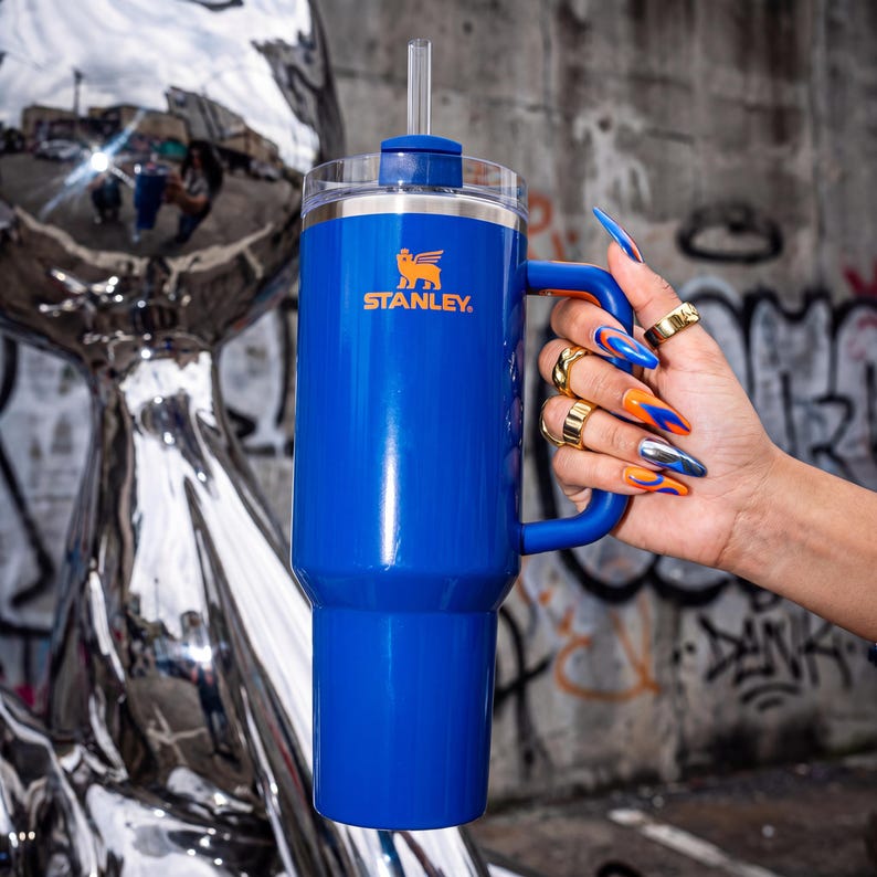 Pode incluir: Um copo Stanley azul com al&ccedil;a e canudo. O logotipo da Stanley &eacute; laranja. O copo &eacute; segurado em frente a uma parede coberta de grafite. A pessoa que segura o copo tem unhas longas e coloridas e an&eacute;is dourados.