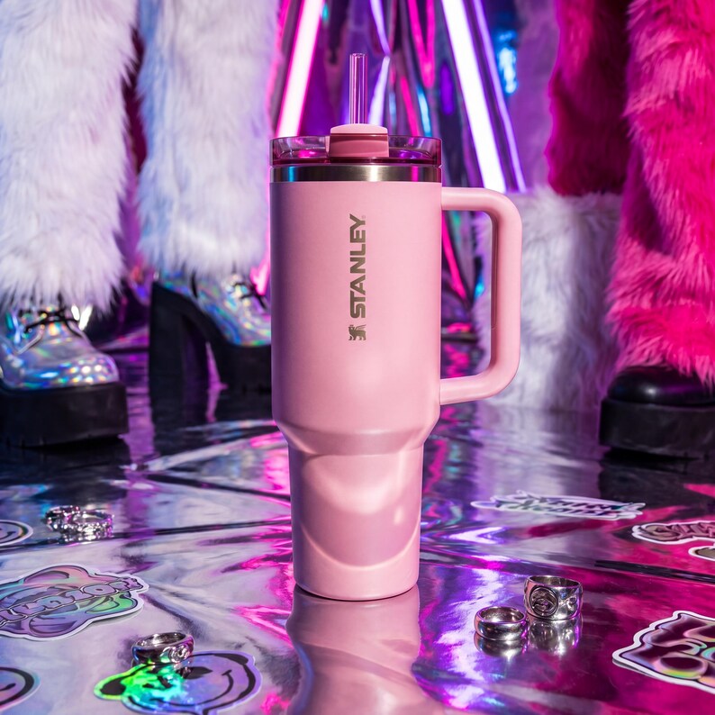 May include: A pink Stanley tumbler with a handle and straw. The tumbler has the Stanley logo printed on the side. The background features a shiny, reflective surface and people wearing platform shoes and furry clothing.
