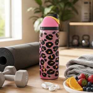 May include: Pink water bottle with a black and orange leopard print design. The bottle has a black lid with a pink top and yellow accent. The word "owala" is printed on the bottle. Other items in the image include a yoga mat, dumbbells, and fruit.