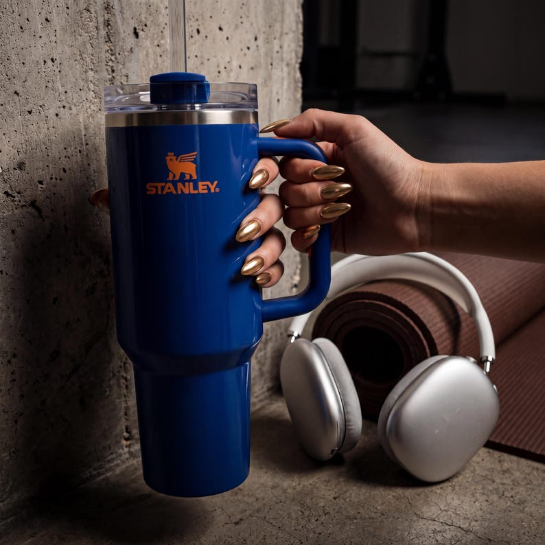 Pode incluir: Um copo Stanley azul com al&ccedil;a, tampa transparente e canudo. O logotipo da Stanley &eacute; impresso em laranja. Fones de ouvido prateados e um tapete de ioga marrom enrolado est&atilde;o ao fundo.