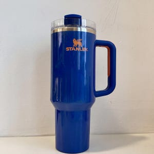 Pode incluir: Uma caneca de viagem Stanley azul com uma al&ccedil;a e tampa transparente. A caneca tem o logotipo Stanley em laranja. A caneca foi projetada para manter as bebidas quentes ou frias. A caneca tem aproximadamente 23 cm de altura.