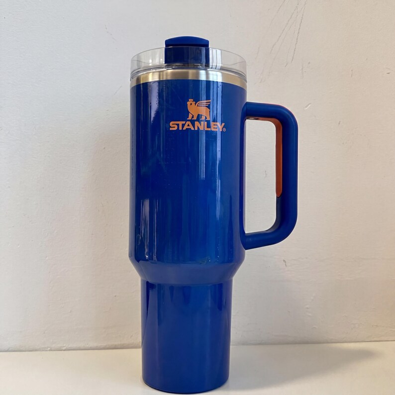 Pode incluir: Uma caneca de viagem Stanley azul com uma al&ccedil;a e tampa transparente. A caneca tem o logotipo Stanley em laranja. A caneca foi projetada para manter as bebidas quentes ou frias. A caneca tem aproximadamente 23 cm de altura.
