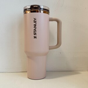 May include: A light pink Stanley travel mug with a handle. The mug has a clear lid and a gold-colored rim. The Stanley logo is printed vertically on the side of the mug.