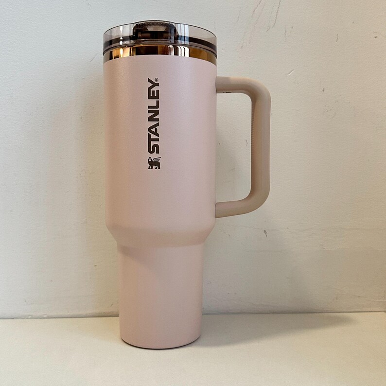 May include: A light pink Stanley travel mug with a handle. The mug has a clear lid and a gold-colored rim. The Stanley logo is printed vertically on the side of the mug.