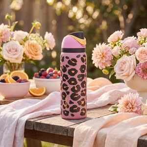 May include: A pink water bottle featuring a black and orange leopard print design. The bottle has a black lid with a yellow handle. The bottle is on a wooden table, with flowers and fruit in the background.
