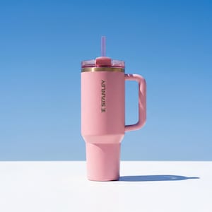 May include: A pink Stanley tumbler with a handle and a clear lid, featuring a straw. The tumbler has a gold rim and the Stanley logo in gold. The background is a gradient of blue and white.