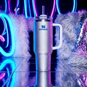 以下が含まれることがあります： 白いハンドルと蓋、透明なストローが付いた、シルバーのスタンレータンブラー。スタンレーのロゴは青色です。タンブラーは、背景にネオンライトのある反射面の上にあります。
