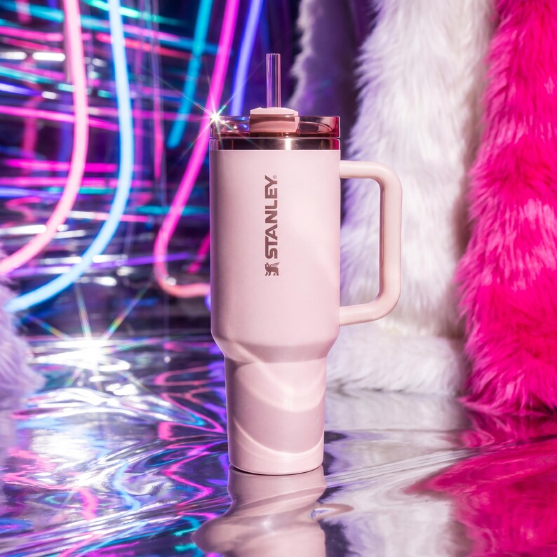 May include: A light pink Stanley tumbler with a handle and a straw. The tumbler has a silver rim and the Stanley logo. The background features colorful neon lights and fluffy pink and white fabric.