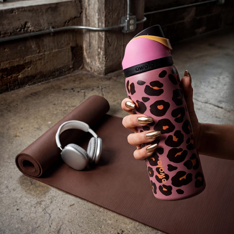 May include: A pink and black leopard print water bottle with a black lid and a yellow accent. The bottle is held by a person with gold fingernails. A brown yoga mat and white headphones are in the background.