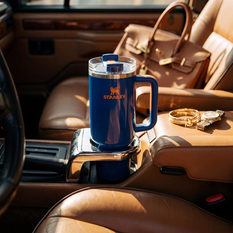 Pode incluir: Uma caneca Stanley azul isolada com uma al&ccedil;a, em um porta-copos de carro. A caneca tem uma tampa transparente e o logotipo Stanley em laranja. Uma bolsa de couro marrom, pulseiras de ouro e um rel&oacute;gio est&atilde;o no fundo.