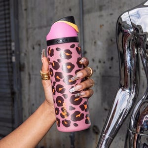 May include: A pink water bottle featuring a leopard print pattern. The bottle has a black and yellow lid and the word "owala" printed on the side. The bottle is held by a hand adorned with gold rings.