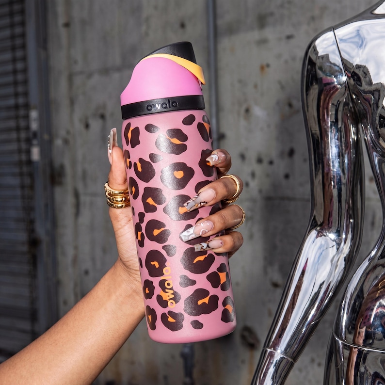May include: A pink water bottle featuring a leopard print pattern. The bottle has a black and yellow lid and the word "owala" printed on the side. The bottle is held by a hand adorned with gold rings.