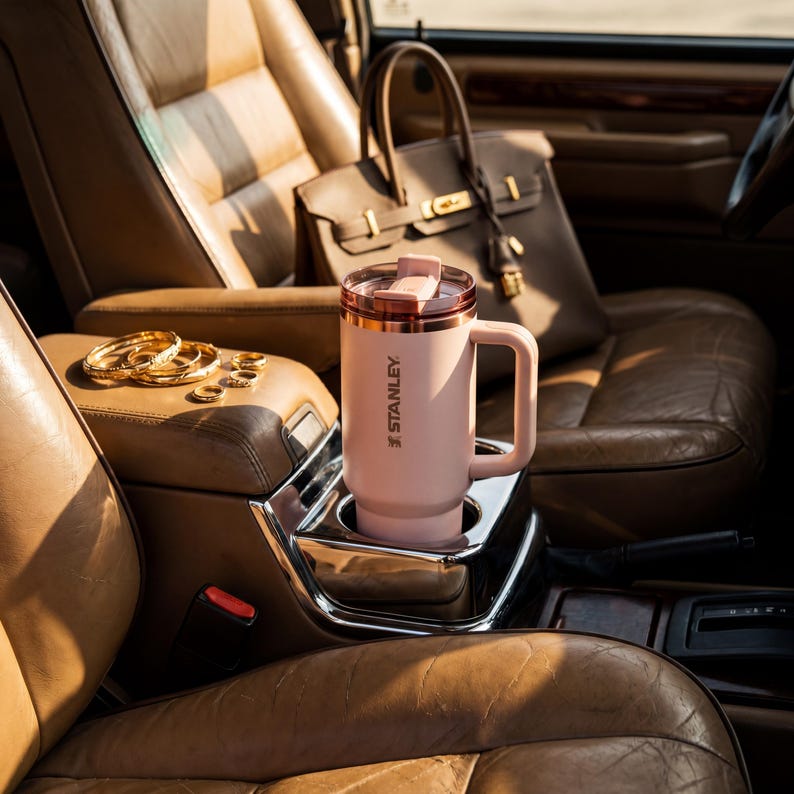 May include: A pink Stanley travel mug with a handle and a rose gold rim sits in a car's cup holder. The mug is next to a tan leather handbag and gold jewelry. The car's interior is brown leather.