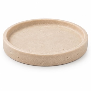 Puede incluir: Una bandeja circular de color beige con una superficie porosa y texturizada. La bandeja presenta un borde ligeramente elevado y una base plana y hundida, mostrando un aspecto natural similar a la piedra con pequeñas hendiduras.